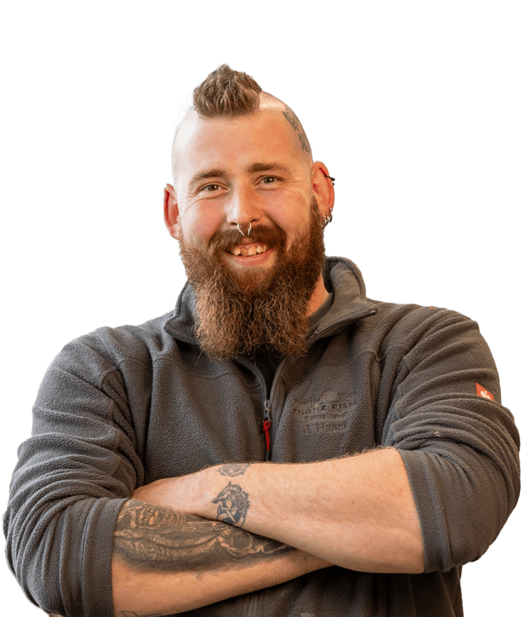 Mann mit Vollbart, hochgestecktem Haar und Tattoos auf dem Unterarm trägt eine Fleecejacke und hat die Arme vor der Brust verschränkt