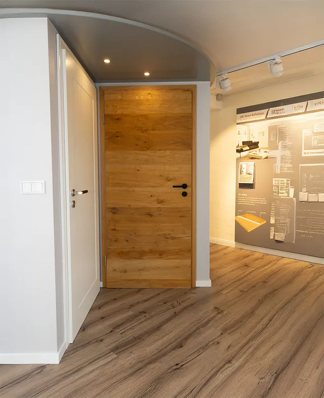 Innenraum mit zwei Türen, eine weiße Tür links und eine Holztür mit schwarzem Griff in der Mitte, heller Bodenbelag und beleuchtete Wand mit Informationspaneel rechts