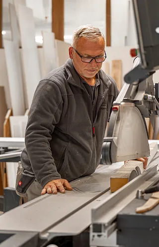 Mann in grauer Arbeitsjacke bedient eine Tischkreissäge in einer Schreinerei mit Holzplatten im Hintergrund