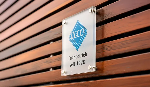 Glasplatte mit blauem VEKA-Logo und Schriftzug 'Fachbetrieb seit 1975' an einer Holzfassade befestigt