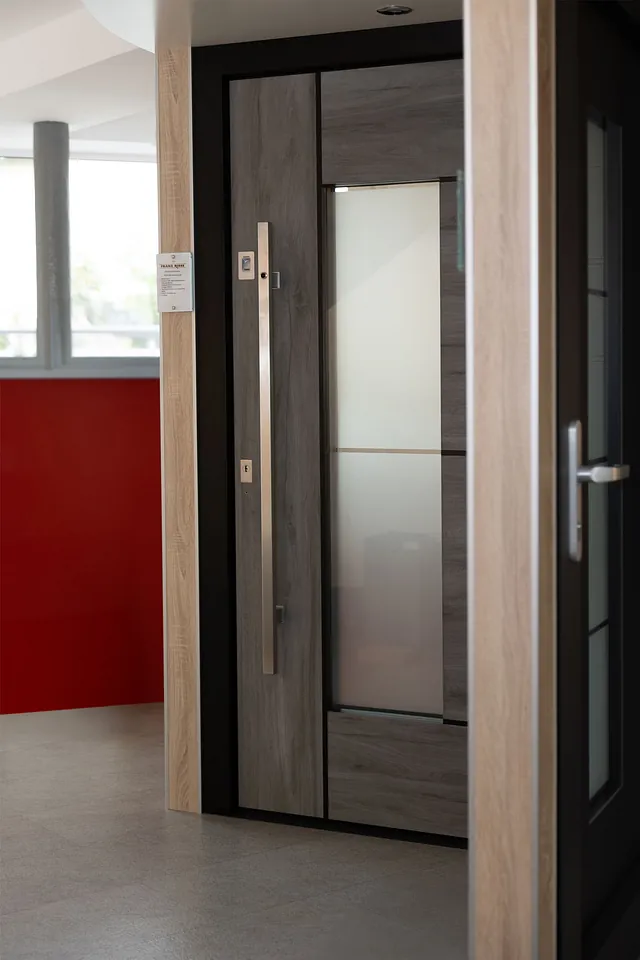 Moderner Hauseingang mit breiter Holztür, vertikalem Griff und seitlichem Fenster in einem Innenraum mit roter Wand im Hintergrund