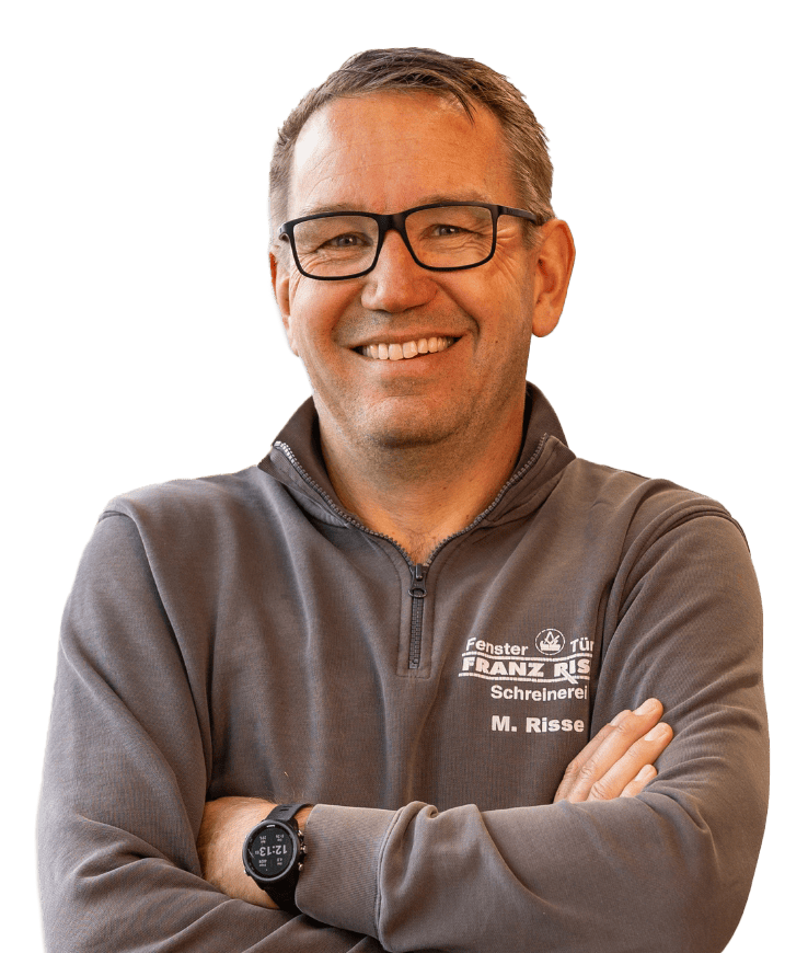 Mann mit verschränkten Armen trägt eine graue Arbeitsjacke mit der Aufschrift 'Fenster Tür FRANZ RISSE Schreinerei M. Risse' und eine schwarze Armbanduhr.